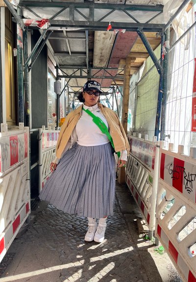 Chaqueta marrón claro sobre una camisa blanca, bolso cruzado verde, falda a cuadros en blanco y negro, zapatillas blancas y una gorra negra en un área de construcción.