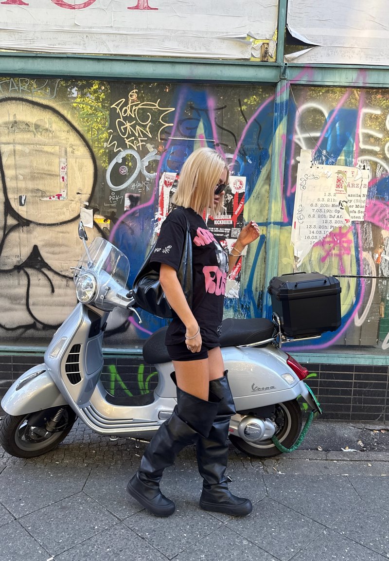 Scooter cinza com um para-brisas transparente estacionado junto a uma parede de graffiti colorido. Usando uma blusa preta oversized, calções curtos e botas altas pretas.