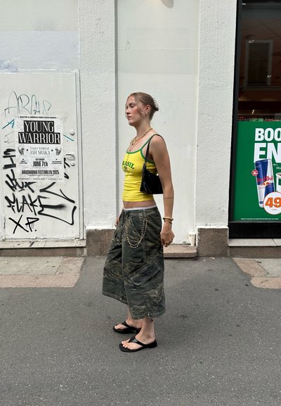 Top de tirantes ajustado amarillo con letras verdes, pantalones cargo de camuflaje con bolsillos, bolso negro y chanclas negras, todo usado en una calle de la ciudad.