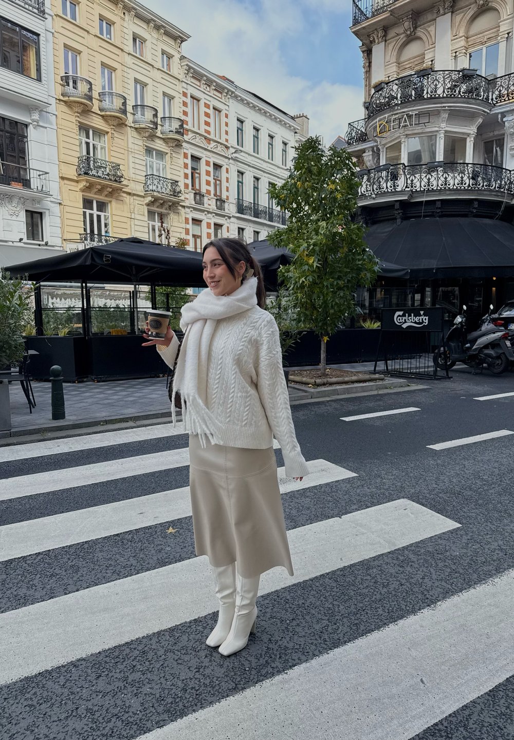 Suéter blanco tejido con patrones de cable, falda de cuero beige y botas blancas hasta la rodilla. Sosteniendo una taza de café cerca de un paso de peatones.