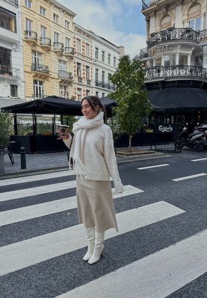 Suéter blanco tejido con patrones de cable, falda de cuero beige y botas blancas hasta la rodilla. Sosteniendo una taza de café cerca de un paso de peatones.