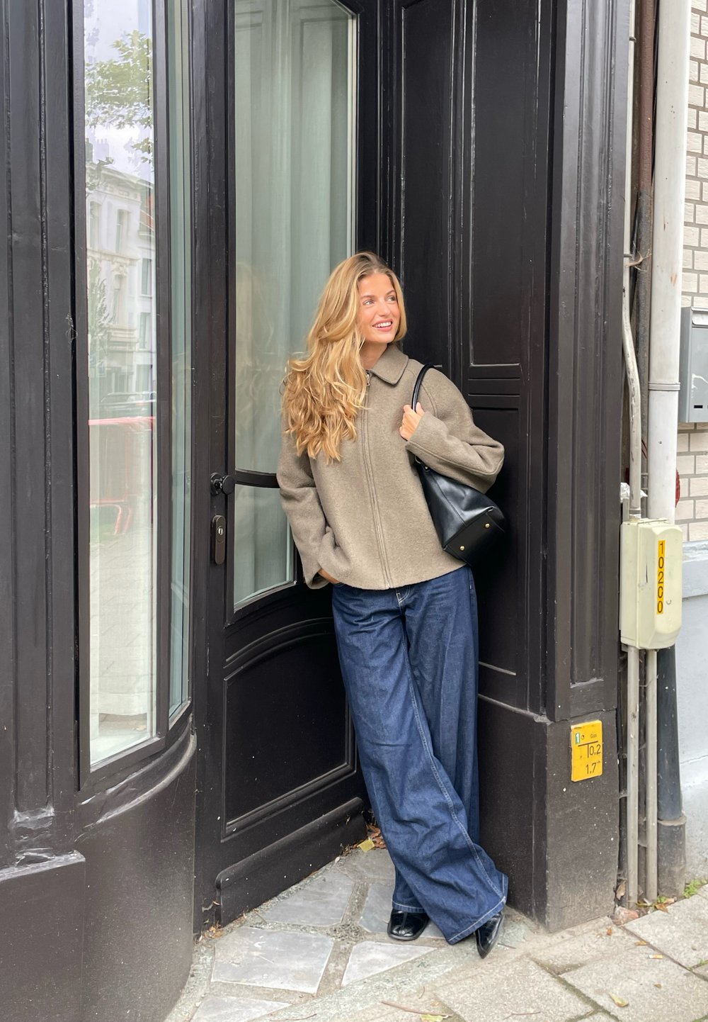 Beige színű, zipzáras dzseki nyakkal, széles lábú kék farmernadrággal és fekete válltáskával viselve. Egy üvegajtó mellett áll.