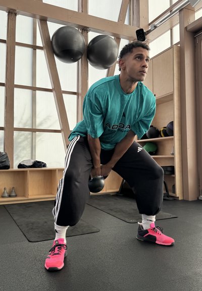 Kettlebell negra, camiseta de gran tamaño color turquesa, pantalones negros con rayas blancas y zapatillas brillantes de color rosa. Entorno de gimnasio con equipo de ejercicio.
