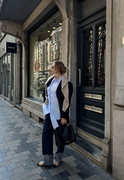 Blazer negro sobre camisa blanca, jeans oscuros con puños de mezclilla, bolso negro y zapatos. Contrastes de textura entre los materiales; fondo de acera y tienda.