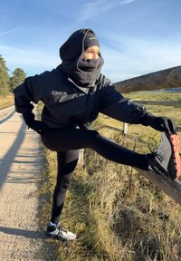 Giacca antivento nera e guanti, leggings neri e scarpe da corsa grigie con dettagli rossi. La persona sta allungando una gamba appoggiandola a un palo di legno.