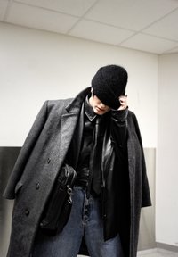 Manteau gris en laine croisé porté par-dessus une veste en cuir noire, cravate foncée et jean en denim bleu. Chapeau tricoté noir et sac en cuir porté en bandoulière visibles.