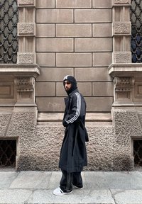Homme portant un long manteau noir, un bonnet en maille, des lunettes de soleil et des baskets blanches, marchant devant un bâtiment en pierre texturée avec des fenêtres décoratives.