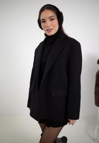 Musta ylipitkä bleiseri, jossa on jämäkät hartiat, yksittäinen napitus ja läppätaskut, päällä musta poolopaita ja hame.