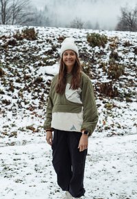 Grønn og beige quiltet jakke med høy krage, oversized design og logo-patch; kombinert med svarte, løstsittende bukser og hvit lue.