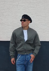 Casaco bomber verde com zip, t-shirt branca por baixo, jeans azuis com detalhe em corrente, boina preta e óculos de sol. Fundo de parede texturizada.