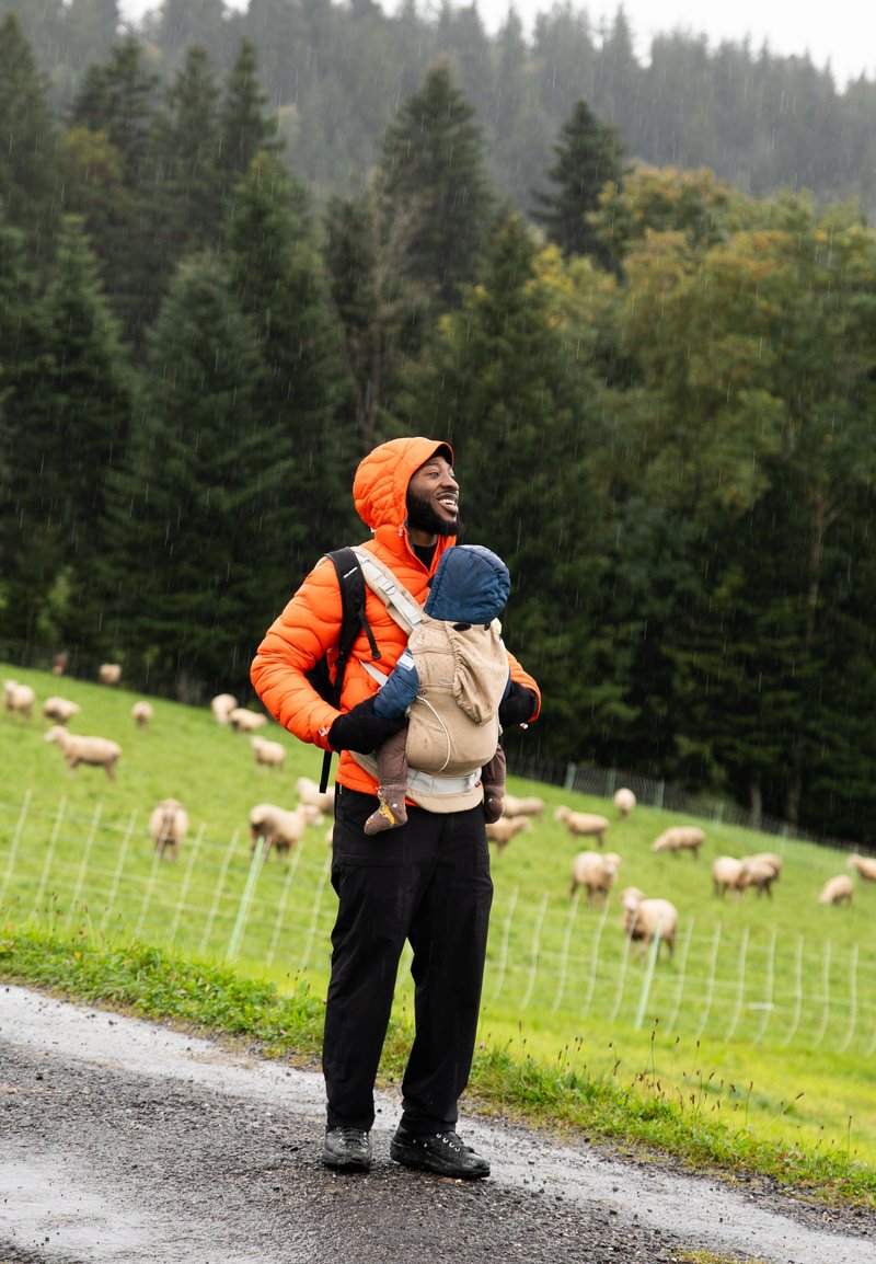 Homme en veste orange porte un bébé dans un porte-bébé, se tenant sur un chemin humide près d'un champ vert avec des moutons paissant sous une légère pluie.