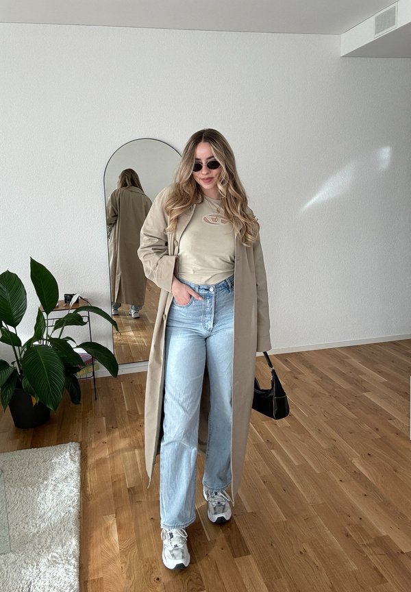 Beige trenčkot přes světle béžové tričko, modré džíny a tenisky. Černé sluneční brýle, černá kabelka a zelená rostlina na pozadí.