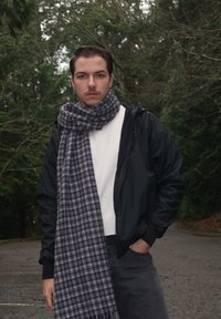 Veste noire légère par-dessus un pull blanc, assortie à une écharpe à carreaux bleu et gris. Pantalon gris et fond de forêt flou.