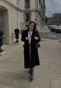 Czarny długi płaszcz noszony na białej bluzce, w zestawieniu z beżową spódnicą i czarnymi kozakami do kolan. Niesie małą czarną torebkę. Ulica miasta.
