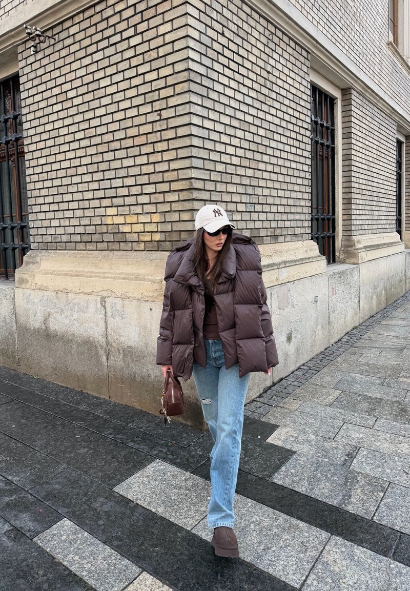 Femme portant une doudoune marron, un jean clair, une casquette blanche et des lunettes de soleil, marchant le long d'un coin de rue pavée en ville, tenant un sac à main marron.