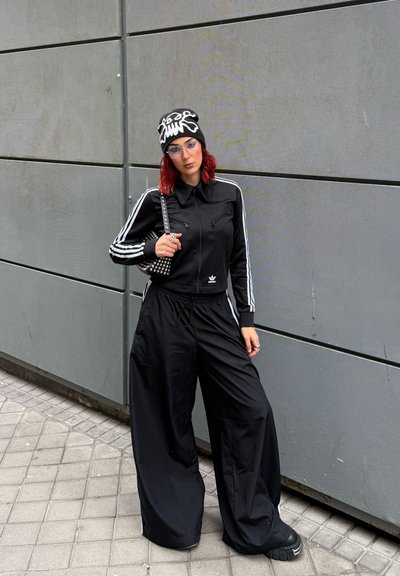 Conjunto deportivo negro que incluye una chaqueta corta con cremallera y rayas blancas, junto con pantalones de pierna ancha. Combinado con un bolso con tachuelas y un gorro con estampado.