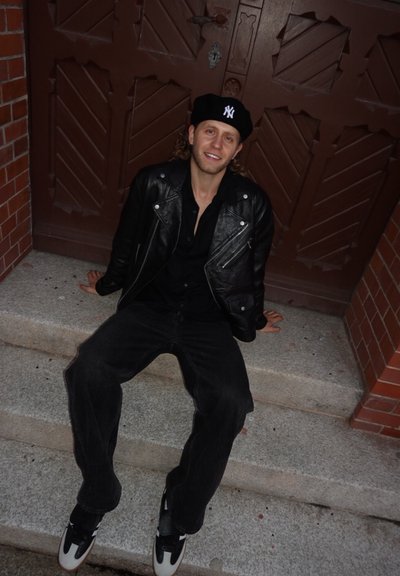 Chaqueta de motociclista de cuero negro, camisa negra, vaqueros negros holgados, zapatillas blancas y negras, con una boina negra con logo, sentado en unas escaleras.