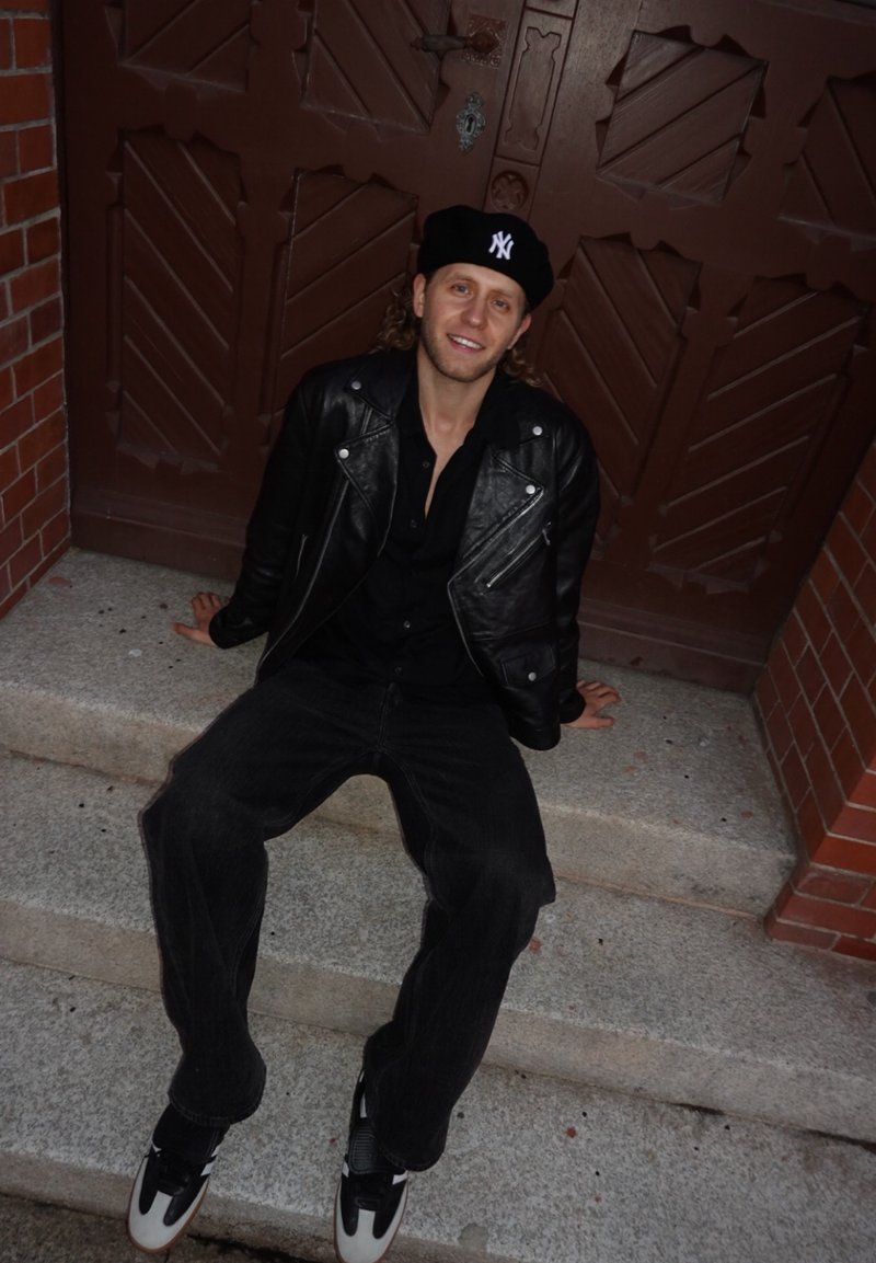 Giacca da motociclista in pelle nera, camicia nera, jeans neri larghi, sneakers bianche e nere, con un berretto nero con un logo, seduto sulle scale.