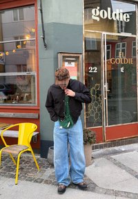 Giacca nera con zip, camicia verde con bottoni sotto, jeans azzurri larghi, berretto marrone a fantasia e scarpe scure in piedi vicino all'ingresso di un caffè.