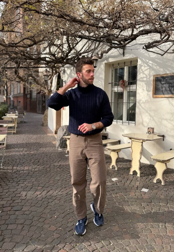 Uomo con maglione blu navy e pantaloni beige in piedi su una strada lastricata di ciottoli fuori da un caffè con tavoli e pergolato con viti senza foglie sopra la testa.