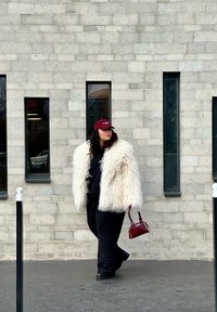 Casaco de pelagem branca e fofa, sweatshirt e calças pretas, boné bordô e uma pequena bolsa bordô. De pé contra uma parede de tijolos cinzas.