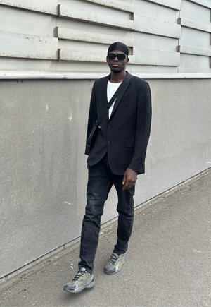 Zwarte blazer over een wit t-shirt, zwarte jeans en grijze sneakers. Donkere zonnebril en een zwarte hoofddoek. Effen gekleurde achtergrond.
