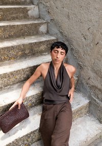 Brązowy jedwabny top na halterze elegancko się układa, w połączeniu z luźnymi brązowymi lnianymi spodniami. W ręce modelki spoczywa pleciona ciemnobrązowa torebka.