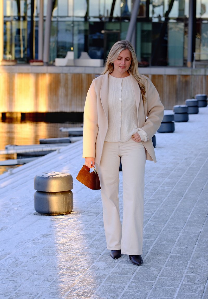 Woman in beige coat and pants walking on snowy pavement near water, holding brown handbag, with modern building in background.
