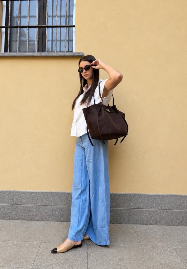 Femme portant des lunettes de soleil, un t-shirt blanc sans manches, un pantalon large bleu et des ballerines beiges, tenant un grand sac à main marron foncé contre un mur jaune.