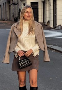 Gebreide kabeltrui in crème, beige geruite blazer, bruine geblokte minirok, gevoerde zwarte handtas, zwarte kniehoge laarzen op straat.