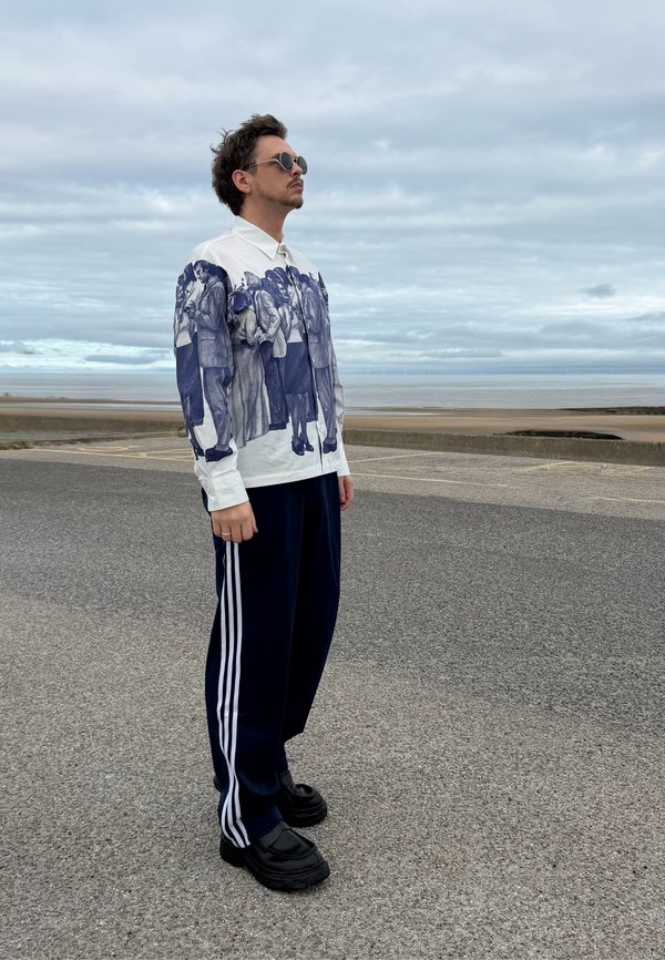 Hombre con gafas de sol, camiseta gráfica de manga larga y pantalones deportivos, de pie en una zona pavimentada cerca de una playa nublada.
