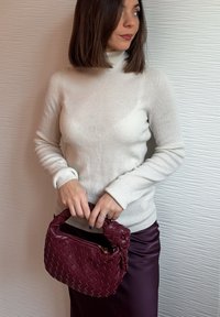 Femme portant un pull à col roulé crème et une jupe violette, tenant un sac à main tressé rouge foncé, regardant vers sa droite devant un mur blanc texturé.