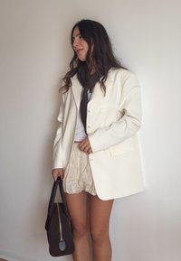 Übergrößen weißer Blazer, weicher Stoff, eine Brusttasche, kombiniert mit einem hellen Oberteil und strukturierten beigen Shorts, sowie einer braunen Handtasche mit goldenen Akzenten.