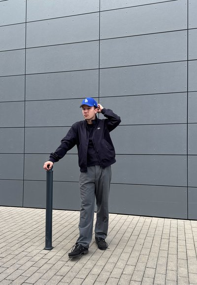 Cazadora bomber negra, pantalones grises, calzado negro; gorra azul con el logo "LA"; de pie junto a un poste gris contra una pared moderna.