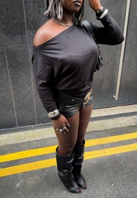 Femme portant un haut noir épaules nues, un short en cuir noir, une large ceinture, des bottes noires ouvertes à l'avant, des bijoux argentés, et portant un sac noir.