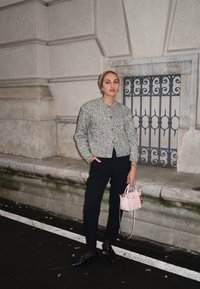 Grijze getextureerde cardigan met gouden knopen, zwarte broek en een lichtroze handtas. De omgeving heeft stenen muren en decoratieve metaalbewerking.