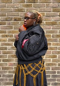 Svart bomberjakke med oransje fôr, med en rød merkelapp på ermet. Kombinert med et svart skjørt pyntet med gule knuter.