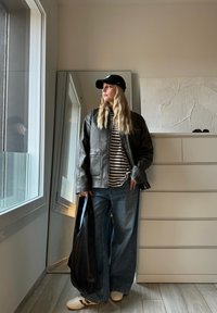 Jaqueta de couro preta, camisa às riscas, jeans azuis largos, mala preta, ténis brancos. Luz refletida da janela e parede texturizada ao fundo.
