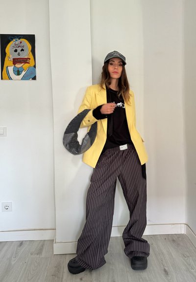 Mujer con blazer amarillo, camiseta gráfica negra, pantalones anchos a rayas, zapatos negros y gorra gris, de pie junto a una pared con pintura colorida.