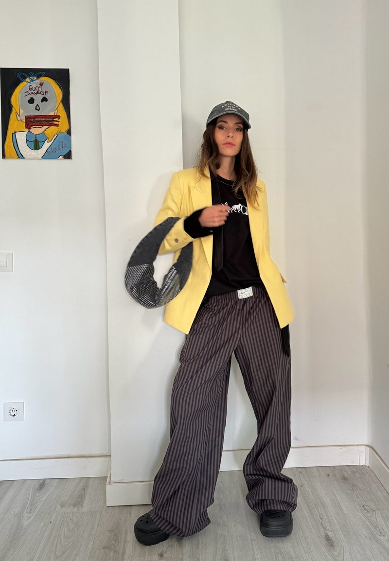 Femme portant un blazer jaune, un t-shirt graphique noir, un pantalon large à rayures, des chaussures noires et une casquette grise, debout à côté d'une peinture murale colorée.