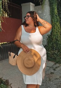 Weißes ärmelloses Kleid aus strukturiertem Stoff mit seitlichem Schlitz. Schwarze Akzente am Ausschnitt, hält einen Strohhut und eine kleine geflochtene Tasche. Grünpflanzen im Hintergrund.