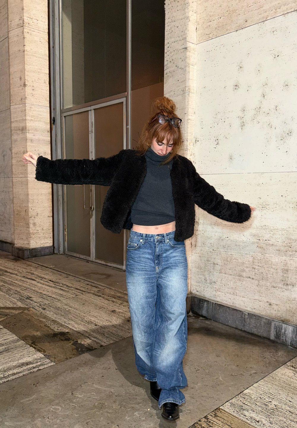 Femme portant des lunettes sur la tête, une veste noire en fourrure, un col roulé court foncé, un jean large bleu et des chaussures noires, debout les bras écartés.