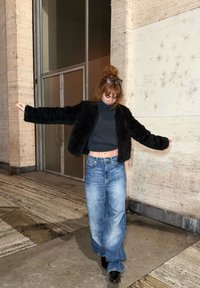 Femme portant des lunettes sur la tête, une veste noire en fourrure, un col roulé court foncé, un jean large bleu et des chaussures noires, debout les bras écartés.