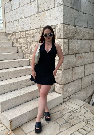 Vestido negro sin mangas con falda acampanada, combinado con sandalias negras de plataforma gruesa. Bolso blanco texturizado y gafas de sol. Fondo de pared de piedra.