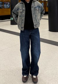 Veste en denim avec une finition usée, sweat à capuche noir en dessous, jeans bleu foncé à coupe décontractée, et baskets marron avec des lacets blancs.