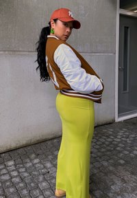 Ruskea ja valkoinen pommitakki, keltainen pitkä hame, vihreät korvakorut ja oranssi lakki; sileät hiukset ponnarilla, seisoo kallioisella päällysteellä.