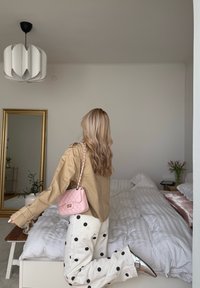 Femme aux longs cheveux blonds portant une veste beige, un pantalon blanc à pois, des chaussures argentées et un sac à main rose, posant dans une chambre minimaliste.