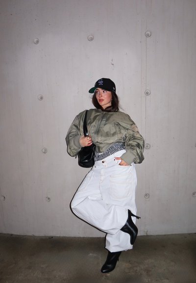 Persona con gorra negra, chaqueta verde holgada, pantalones blancos anchos, botas negras de tacón y llevando un bolso negro de hombro frente a una pared gris.