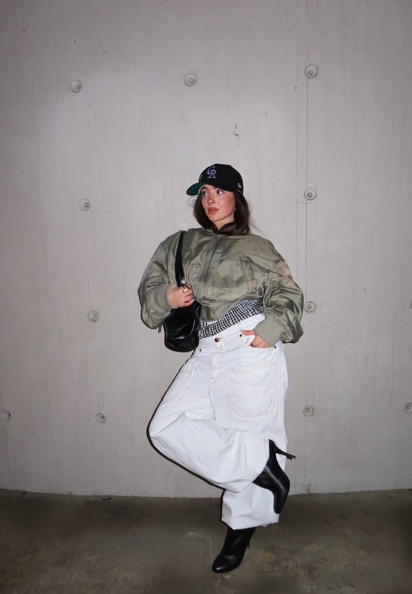Osoba v čiernej čiapke, veľkej zelenej bunde, bielych voľných nohaviciach, čiernych členkových čižmách na podpätku a s čiernou kabelkou cez rameno stojaca pred šedou stenou.
