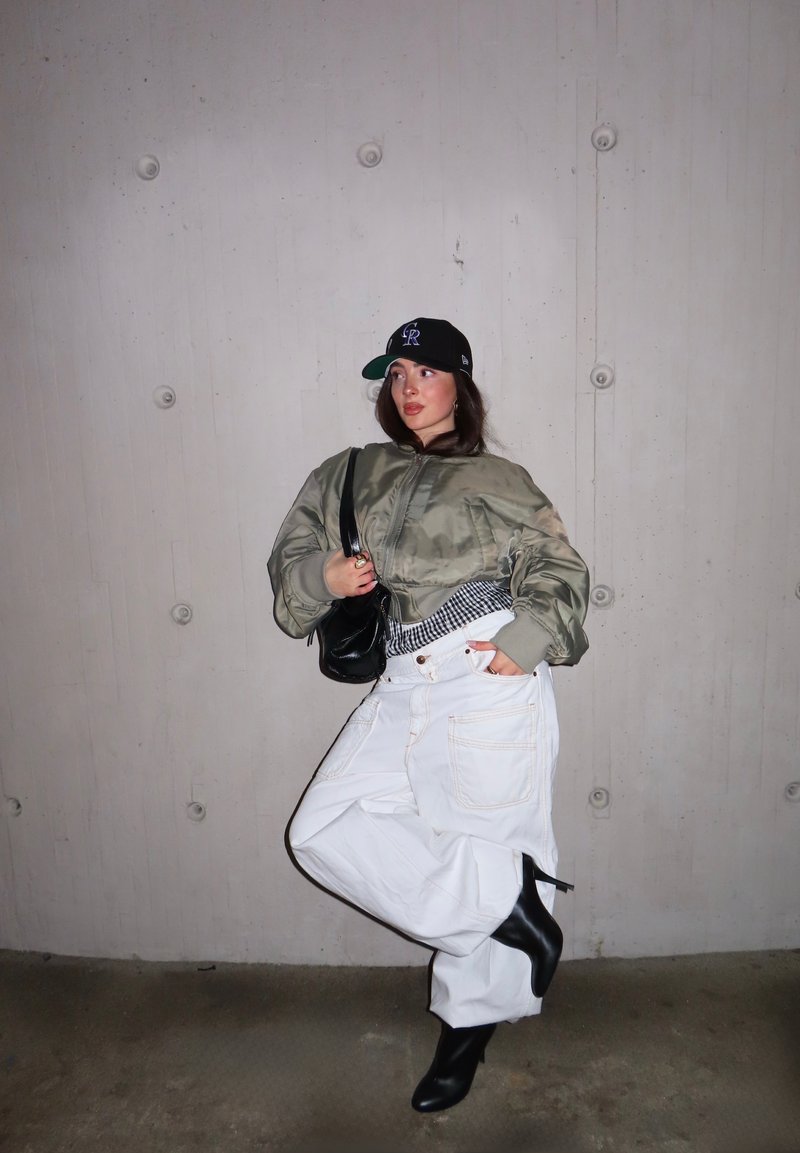 Personne portant une casquette noire, une veste verte surdimensionnée, un pantalon blanc ample, des bottines noires à talons et un sac à bandoulière noir devant un mur gris.